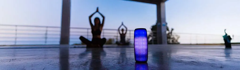 Bluetooth speaker met blauwe verlichting op de voorgrond, met op de achtergrond twee mensen in yogahouding op een open terras bij zonsondergang.