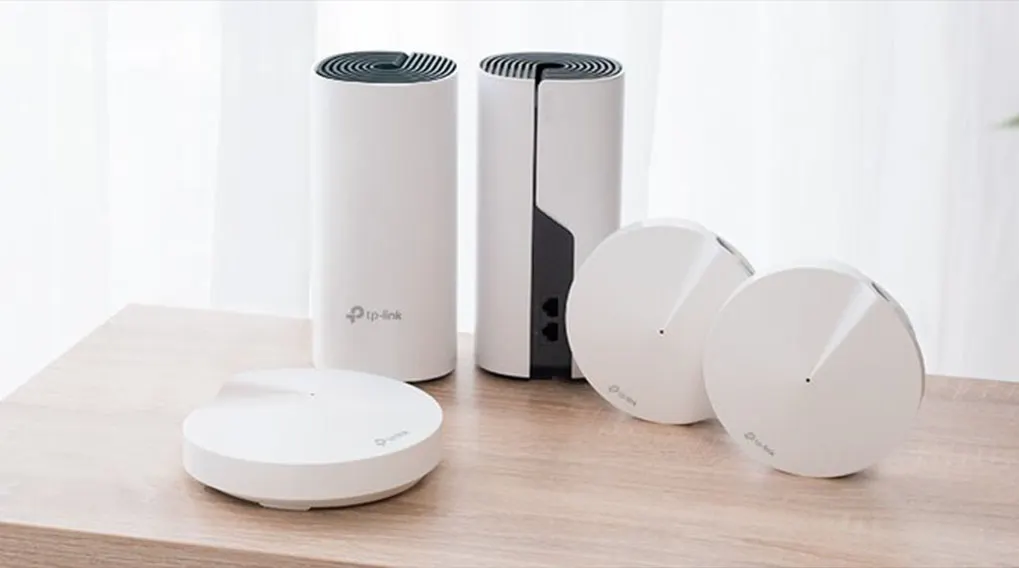 Twee witte, cilindervormige TP-Link-routers en twee platte, ronde routers staan op een houten tafelblad voor een wit gordijn.