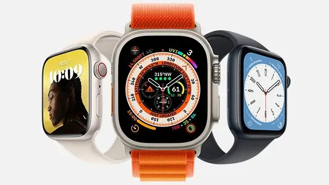 Drie Apple Watches staan op een witte achtergrond. De middelste heeft een oranje band en een scherm met veel wijzers. De andere hebben een lichte en donkere band.
