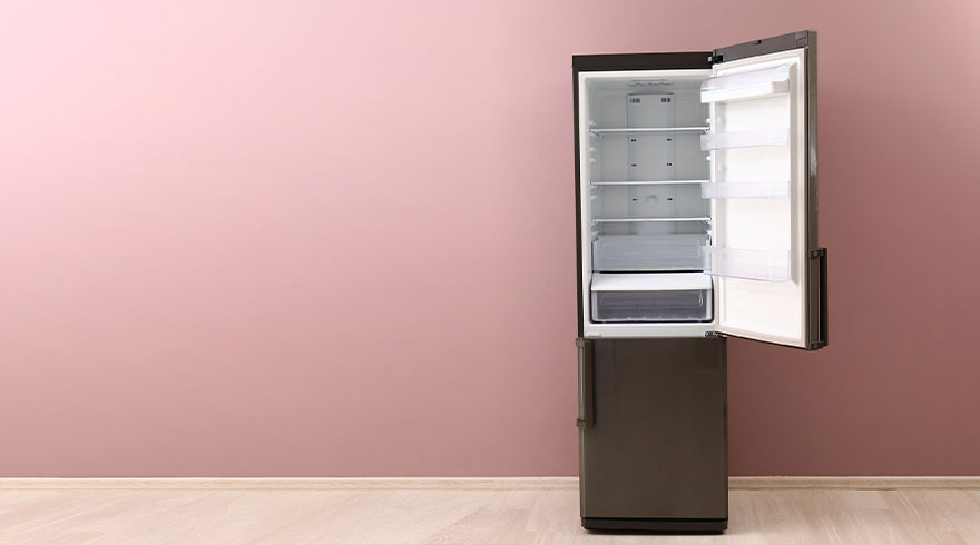 Een openstaande, donkergrijze koelkast staat tegen een roze muur op een houten vloer. De koelkast is leeg met witte planken.