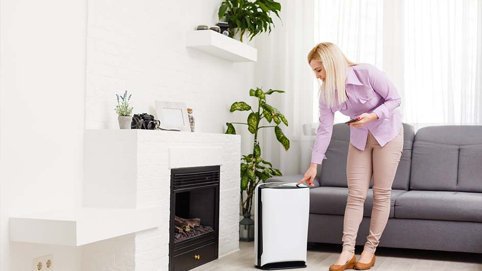 Een vrouw drukt op een witte luchtreiniger naast een grijze bank en witte open haard in een lichte kamer met planten.