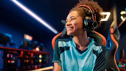 Een glimlachende persoon met een blauw-zwart shirt en een oranje koptelefoon zit in een gamestoel. Neonlichten op de achtergrond.