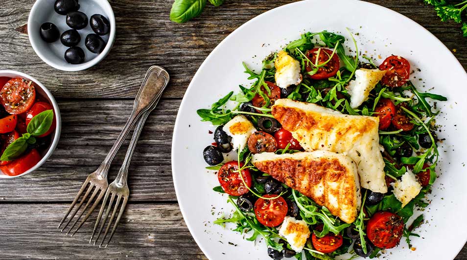 Een bord met salade, gegrilde vis, tomaat en olijven, naast kommen met tomaatjes en olijven, en twee vorken op hout.