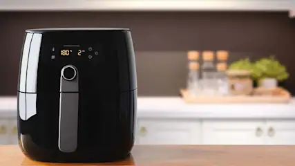 Een zwarte airfryer met digitaal display op een houten aanrecht, met flessen en groen decor op de achtergrond.