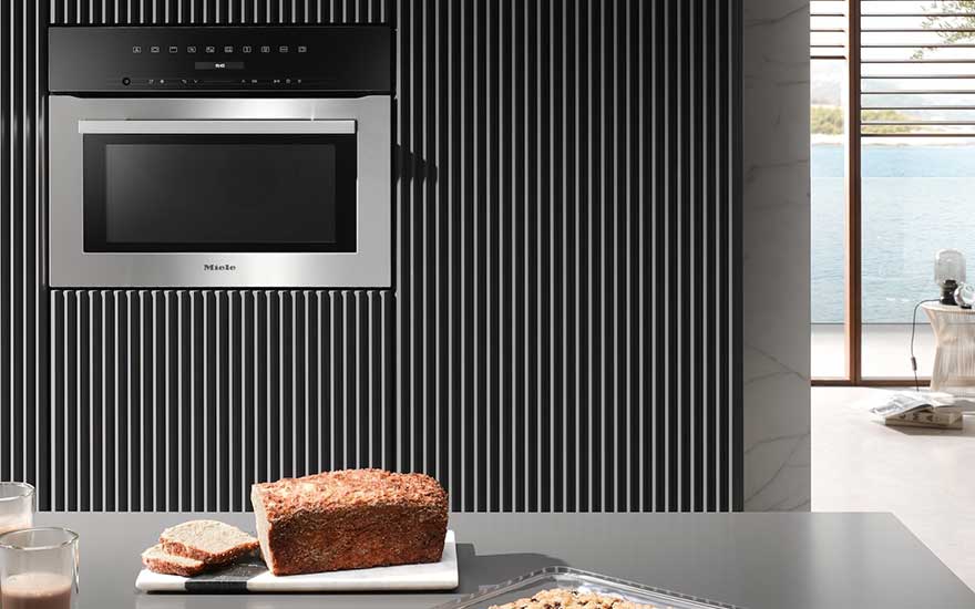 A sleek Miele microwave is built into a modern black striped wall; bread, slices, and drinks rest on a grey worktop in a bright kitchen.