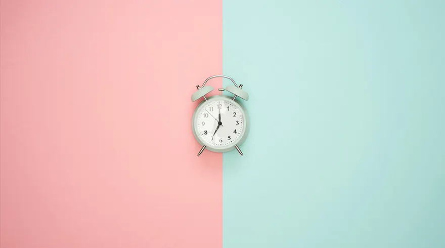 Een wekker staat centraal op een achtergrond die verticaal in roze en blauw is verdeeld.