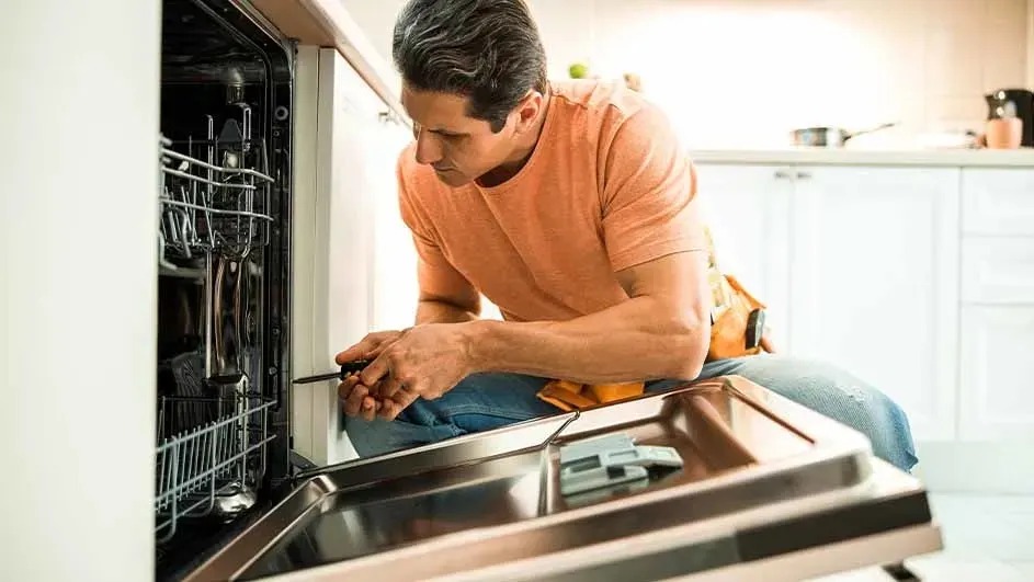 Een man repareert een ingebouwde vaatwasser in een moderne keuken. Het merk van de vaatwasser is niet zichtbaar op de afbeelding.