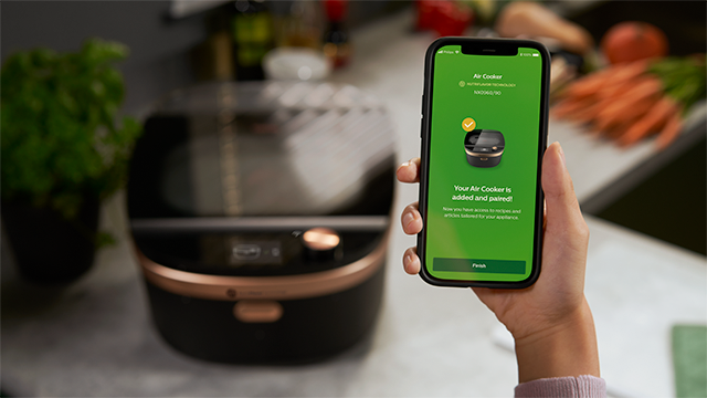 Een hand houdt een telefoon met een groene app naast een zwarte airfryer met koperen accenten. Wortelen liggen op de achtergrond.
