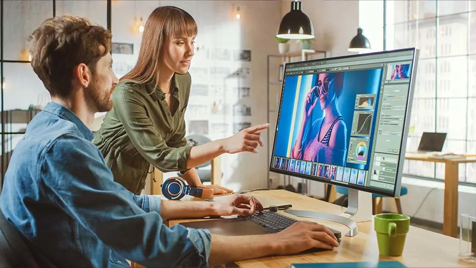 Een man en een vrouw werken samen aan een computer met foto-editing software in een lichte kantoorruimte.