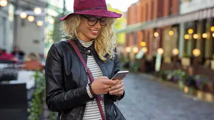 Een jonge blonde vrouw met een bordeaux hoed leest lachend op haar smartphone terwijl ze op een terras in de stad zit.