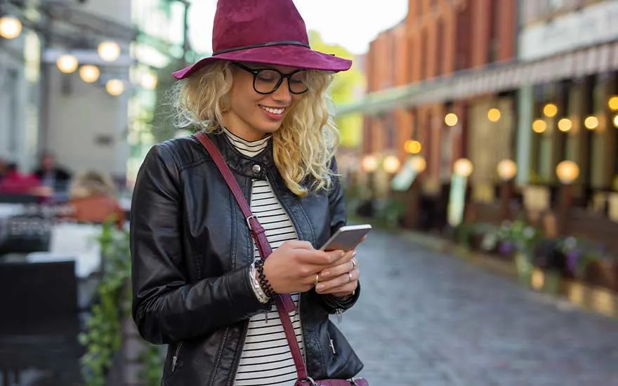 Een jonge blonde vrouw met een bordeaux hoed leest lachend op haar smartphone terwijl ze op een terras in de stad zit.