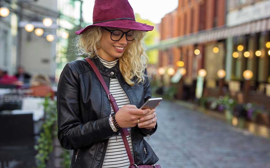 A young blonde woman in a burgundy hat smiles as she reads something on her smartphone while sitting outside in a lively street cafe.