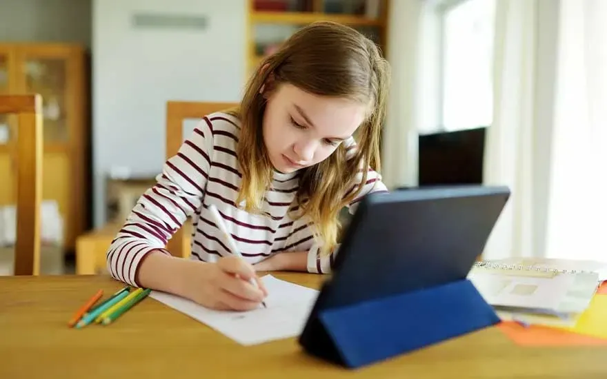 Een meisje met lang haar maakt huiswerk aan een houten tafel, schrijft op papier en gebruikt een tablet met een blauwe hoes. Merken zijn niet zichtbaar.