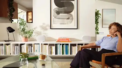 Een vrouw zit in een stoel voor een witte boekenkast met boeken, planten, lamp en kunst aan de muur.