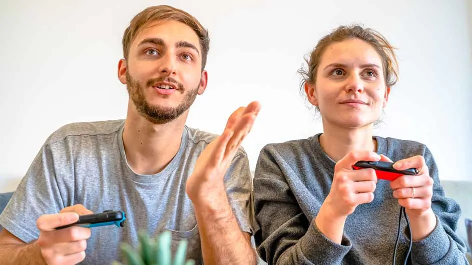 Een man en een vrouw spelen een videospel met rode en blauwe controllers tegen een witte achtergrond.