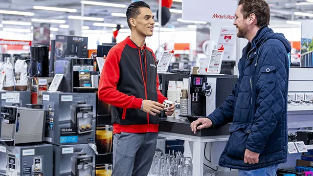 Een verkoper in een rood-zwart vest staat tegenover een klant in een blauwe jas in een winkel met koffiemachines.