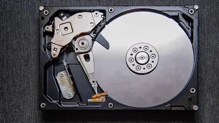 Een bovenaanzicht van een opengewerkte harde schijf met een zilveren schijf en metalen leesarm tegen een donkere achtergrond.