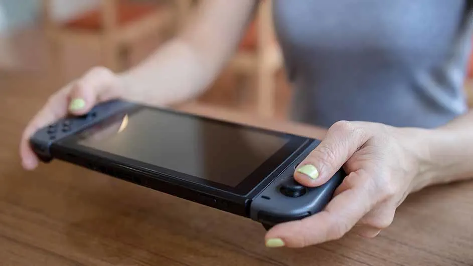Een vrouw houdt een zwarte Nintendo Switch vast boven een houten tafel.