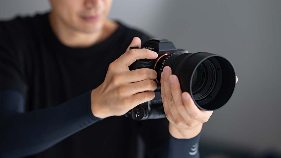 Een persoon in een zwart shirt houdt een zwarte camera met een grote lens tegen een grijze achtergrond.