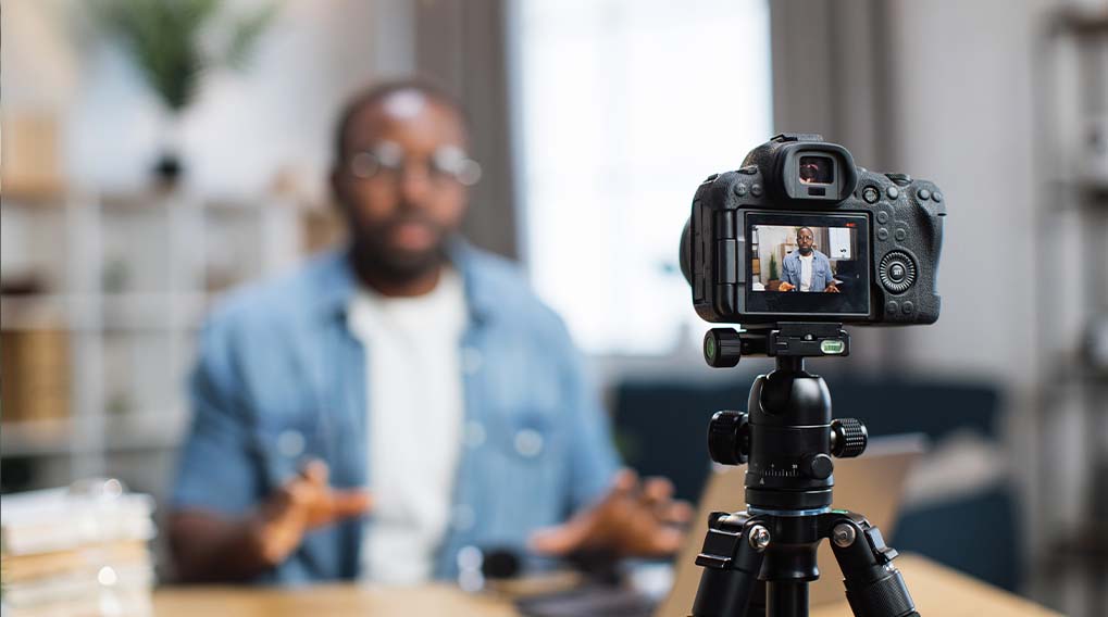 Een zwarte camera op een statief filmt een persoon in een blauw overhemd voor een plankenkast.