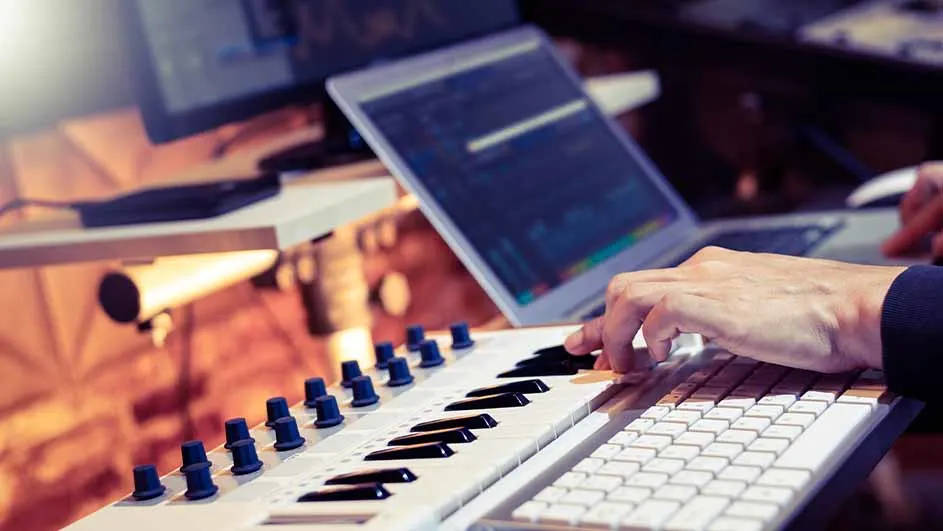 Een hand speelt op een wit keyboard met zwarte toetsen, naast een witte computertoetsenbord, met laptops en monitoren op de achtergrond.