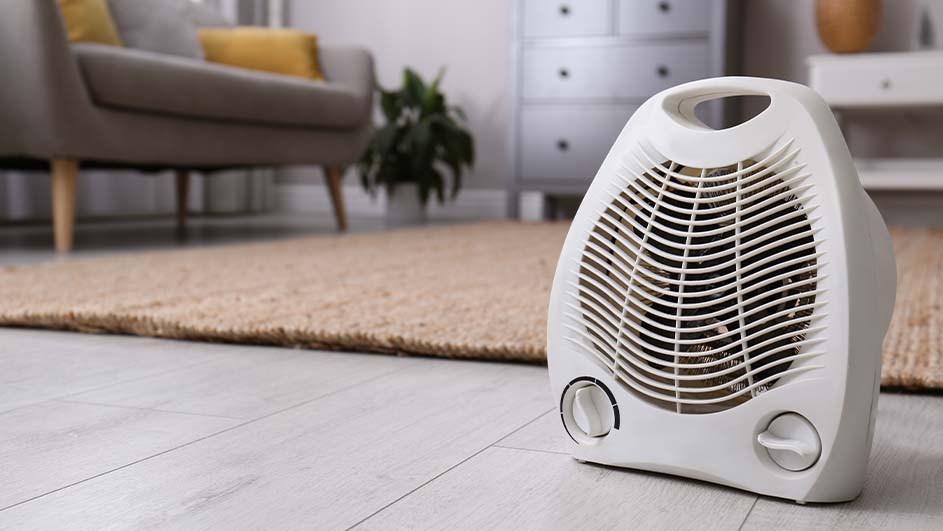 Een witte elektrische kachel staat op een houten vloer, met een beige tapijt, een grijze stoel en planten op de achtergrond.