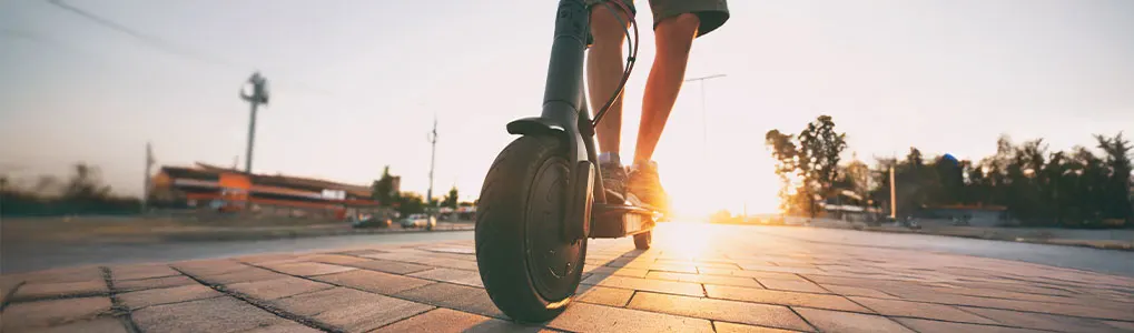A person rides a scooter on a brick path toward a bright sunset. Buildings and trees are in the background.