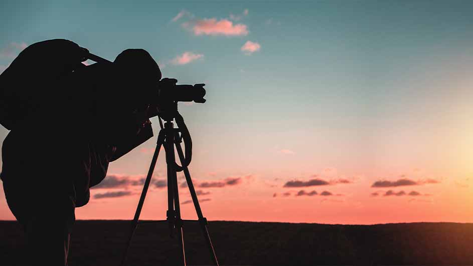 Silhouet van een persoon met een camera op een statief tegen een zonsondergang met een blauwe en oranje hemel.