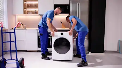Twee mannen in blauwe uniformen tillen een witte wasmachine in een keuken. Een blauwe steekwagen staat links.