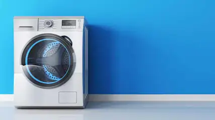 Een witte wasmachine met een zwart deur tegen een blauwe achtergrond en een witte plint.