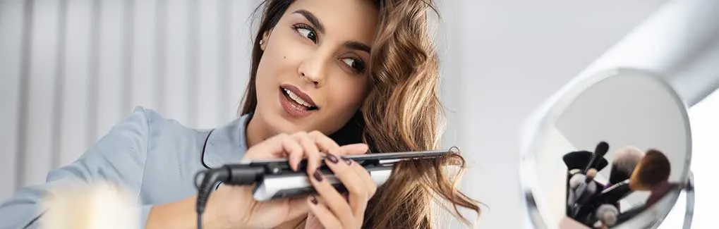 A woman styles her wavy hair with straighteners by a mirror, while make-up brushes are visible in the foreground.
