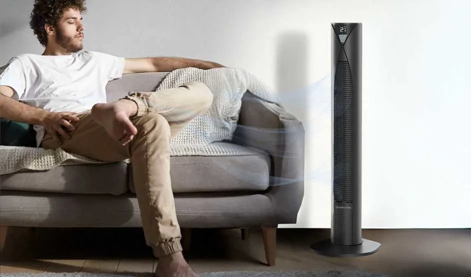 A man relaxes on the sofa beside a slim tower fan; soft air currents suggest coolness in a bright living room.