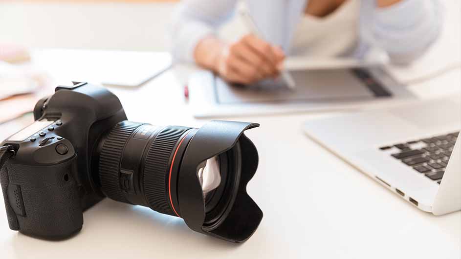 Een zwarte camera met een grote lens ligt op een witte tafel, naast een laptop en een grafisch tablet, met een persoon op de achtergrond.