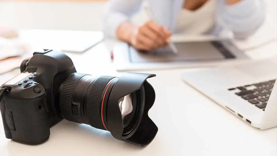 Een zwarte camera met een grote lens ligt op een witte tafel, naast een laptop en een grafisch tablet, met een persoon op de achtergrond.