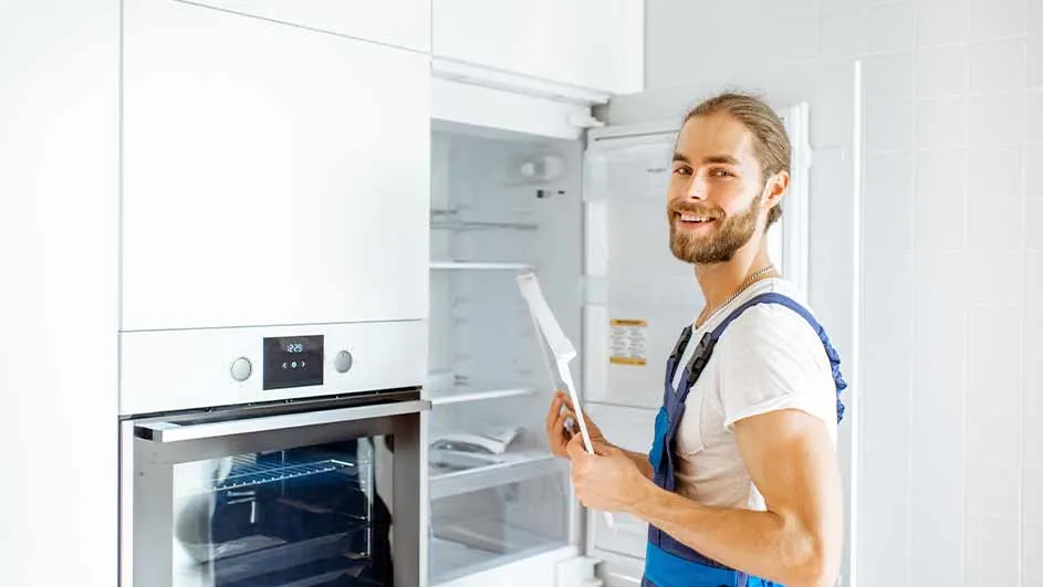 Een man in blauwe overall staat bij een open witte koelkast naast een witte oven in een witte keuken.