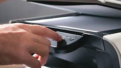 Een hand drukt op de knop van een openstaande grijze en zwarte printer tegen een grijze achtergrond.