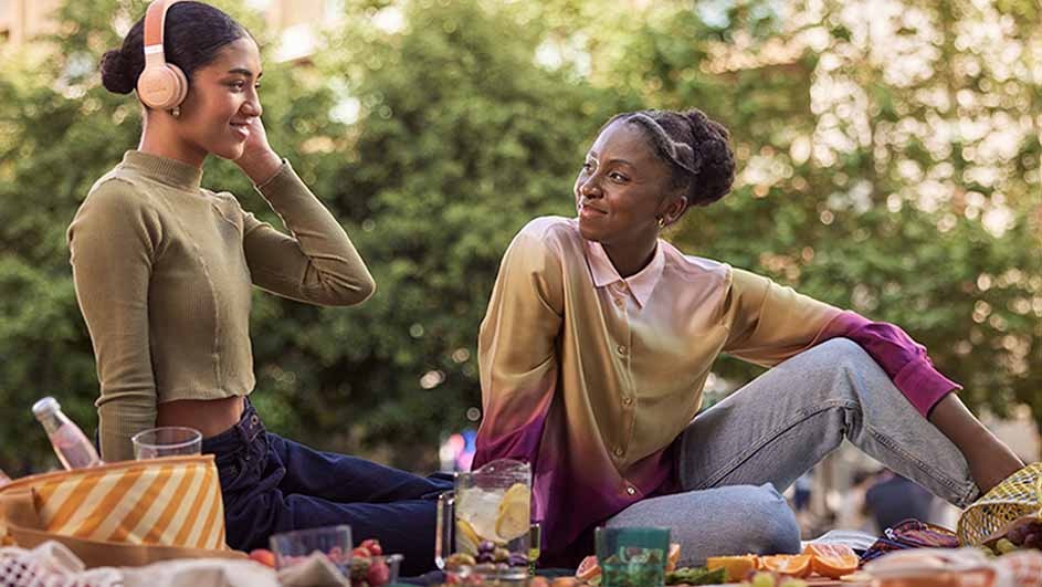 Two women enjoy a picnic with drinks and sliced fruit. One wears headphones. Trees are visible in the background.