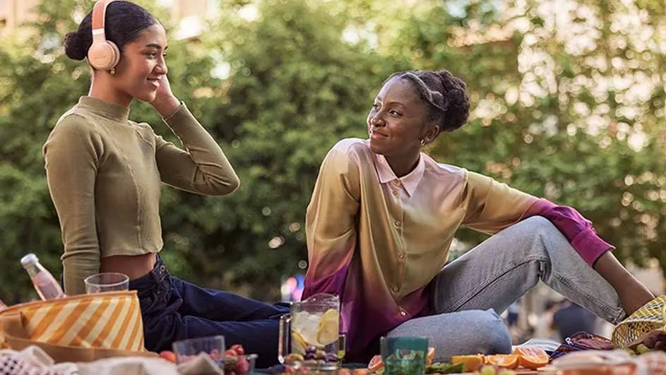 Two women enjoy a picnic with drinks and sliced fruit. One wears headphones. Trees are visible in the background.