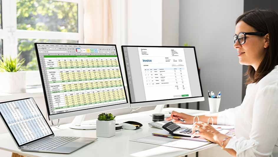 A woman works at a desk with two monitors, a laptop, a calculator, and an invoice. A window is visible in the background.