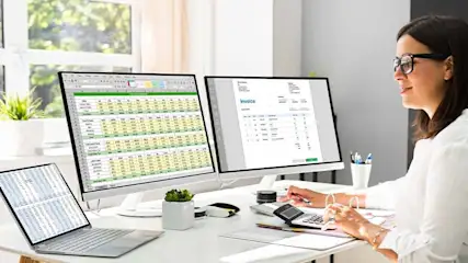 A woman works at a desk with two monitors, a laptop, a calculator, and an invoice. A window is visible in the background.