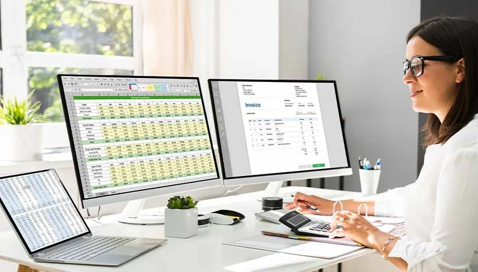 A woman works at a desk with two monitors, a laptop, a calculator, and an invoice. A window is visible in the background.