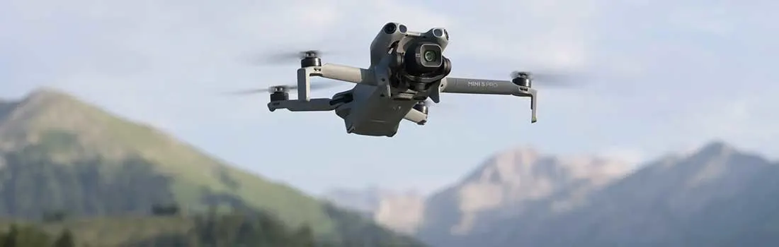 A grey drone hovers mid-air, set against a backdrop of green mountains and blue skies, ready to capture photos.