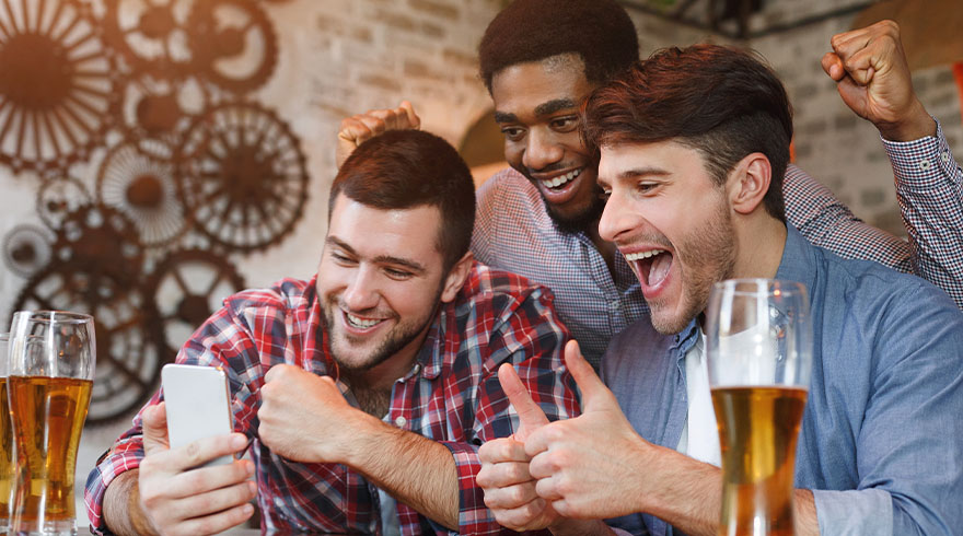 Drie mannen vieren feest met bier. Een man houdt een witte smartphone vast. Op de achtergrond een decoratieve wand met tandwielen.