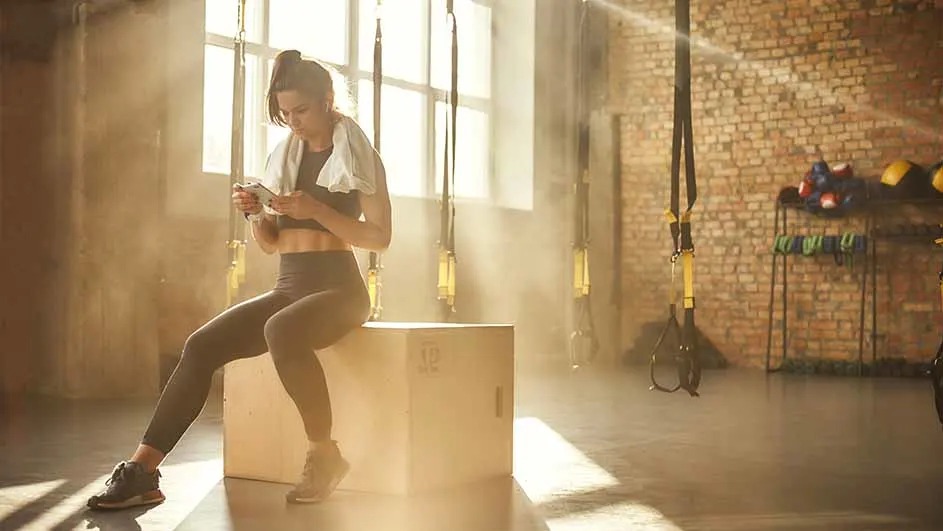 Sportende vrouw zit op een houten plyobox in een zonovergoten gym, handdoek om haar nek, kijkend op haar telefoon tussen de TRX-banden.