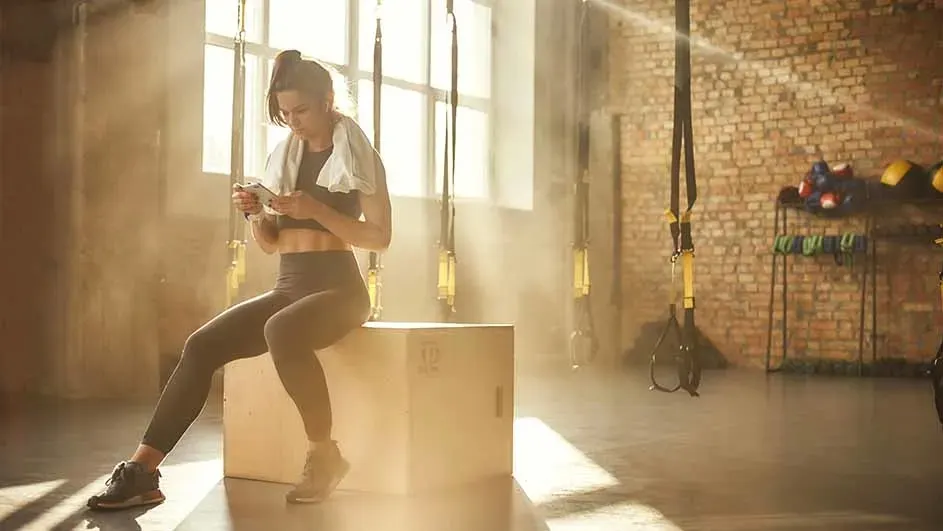 Sportende vrouw zit op een houten plyobox in een zonovergoten gym, handdoek om haar nek, kijkend op haar telefoon tussen de TRX-banden.