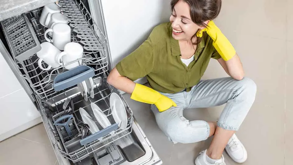 Vrouw met gele handschoenen zit naast een geopende vaatwasser vol witte mokken en borden.