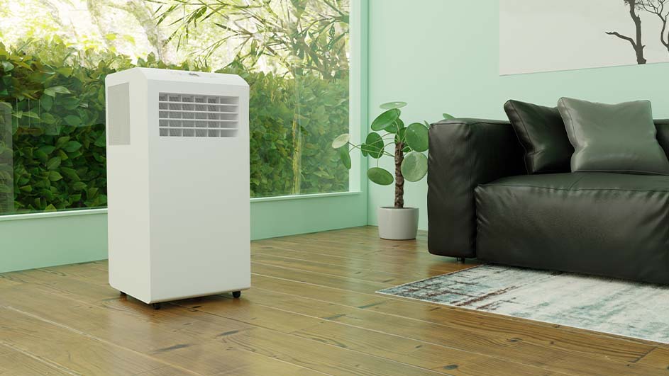 Een witte mobiele airconditioner staat in een kamer met een zwarte bank en een kamerplant.