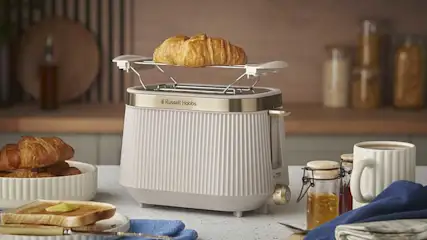 A stylish white Russell Hobbs toaster with warming rack holds a croissant, surrounded by toast with butter, jam jars, a white mug of coffee and breakfast dishes.