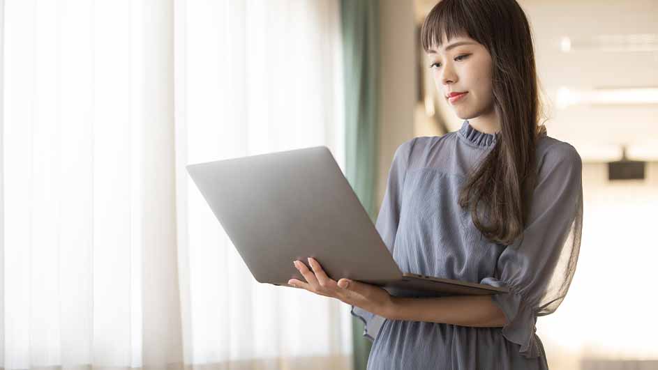 Een vrouw in een grijze jurk staat voor een raam en houdt een grijze laptop vast.