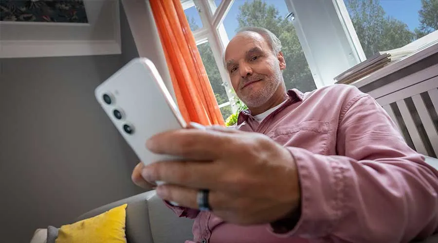 Person in a pink shirt looks at a smartphone by a window with orange curtains, sitting on a sofa with a yellow cushion.